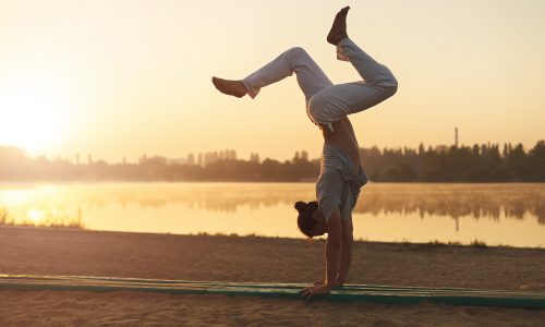 Concept about lifestyle and sport. Capoeira on the beach, near lake one performer, at sunrise. Athletic man in white pants standing on arms upside down while workout in the park in the heart of city.